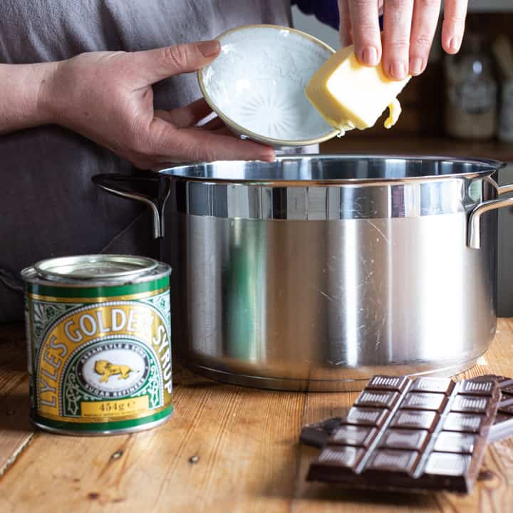 womans hands holding a stick of butter over a large silver saucepan on a wooden kitchen counter surrounded by baking mess