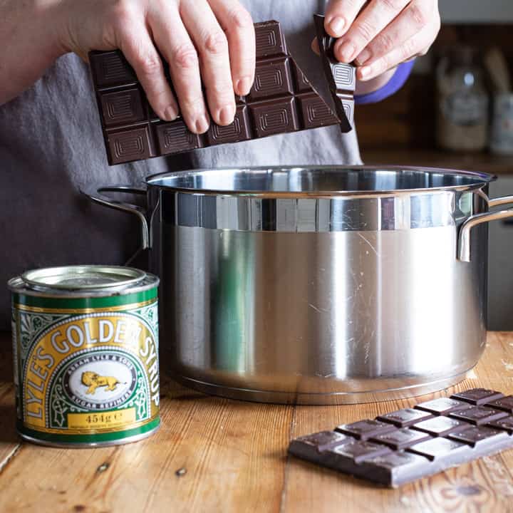 womans hands breaking dark chocolate pieces over a large silver saucepan on a wooden kitchen counter surrounded by baking mess