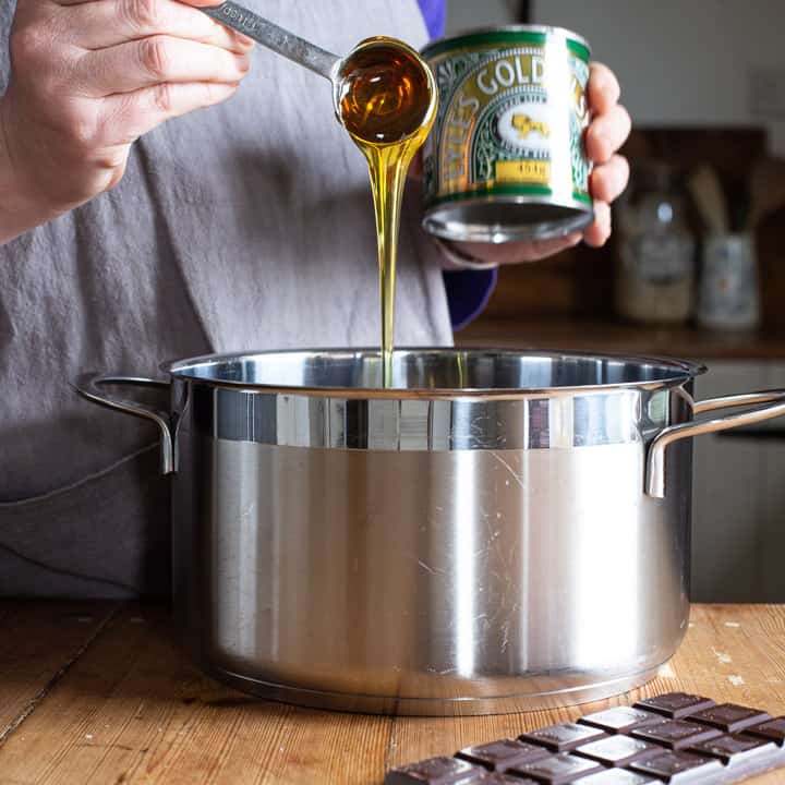 womans hands spooning golden syrup from a green can into a large silver saucepan on a wooden kitchen counter surrounded by baking mess