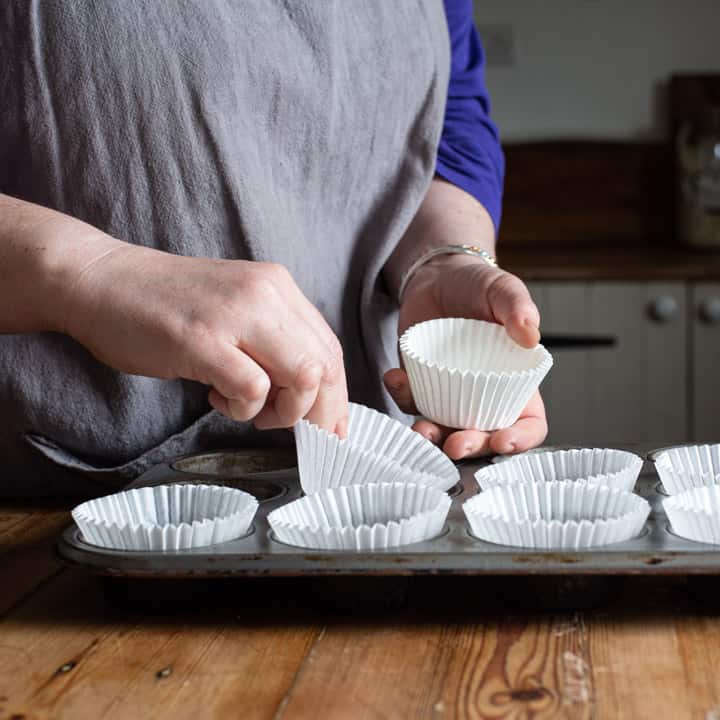 womans hands placing white cupcake cases into a silver cupcake baking tin