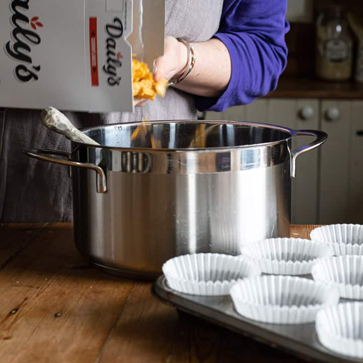 womans hands pouring yellow cornflakes from a white cereal box into a large silver saucepan on a wooden kitchen counter 