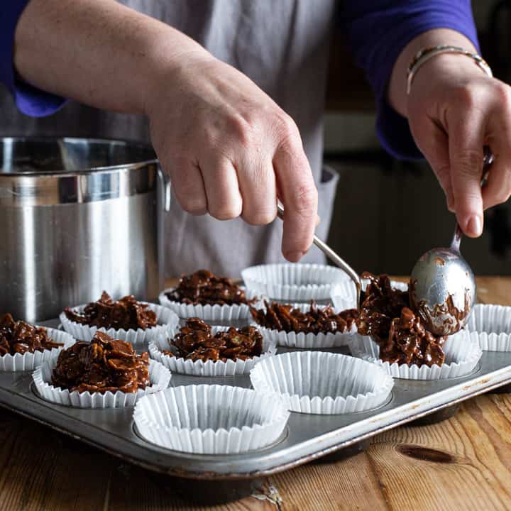 womans hands spooning chocolate covered cornflakes into white paper cupcake cases on a silver tray