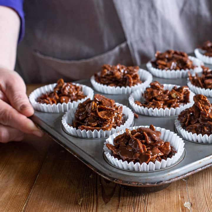 womans hands holding a silver metal tray of white paper cases filled with chocolate covered cornflakes