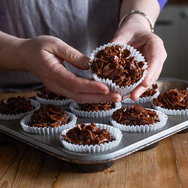 womans hands holding a chocolate cornflake cake over a full tray of cakes