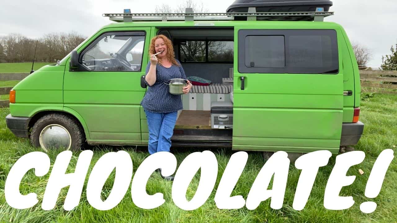 woman with red hair wearing blue clothes stood in front of a bright green campervan licking a a chocolate coated spoon