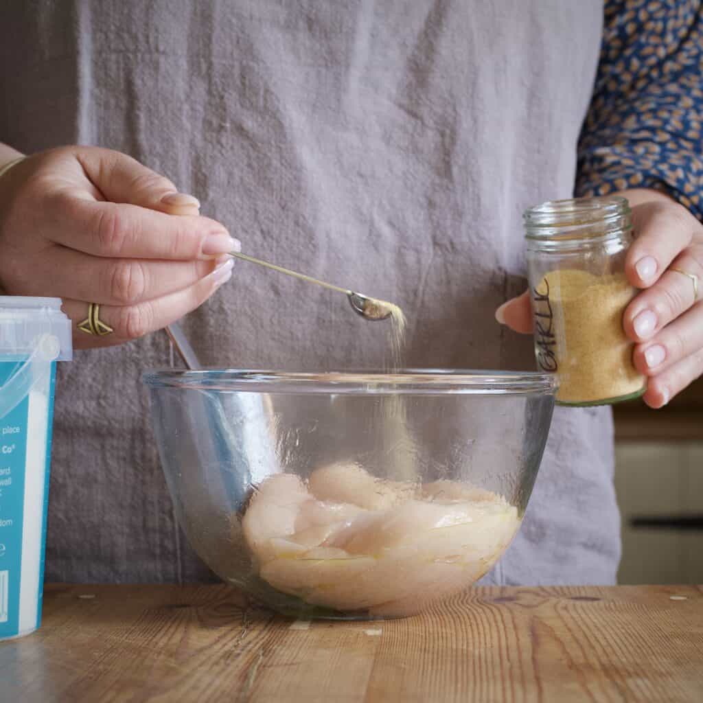 Womans hands spooning garlic powder onto several chicken fillets in a glass mixing bowl on a wooden kitchen counter