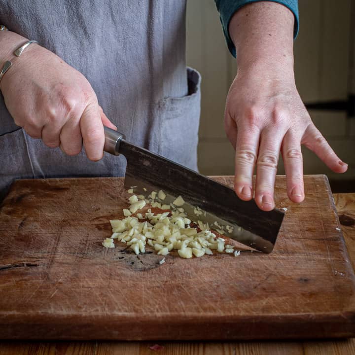 womans hands finely chopping garlic with large Japanese knife on wooden board