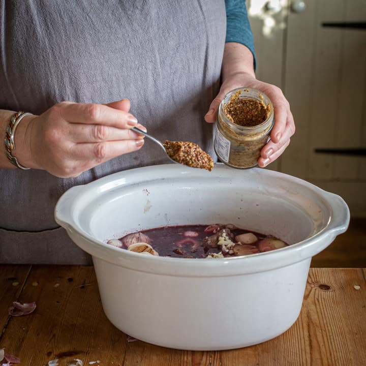 womans hands spooning wholegrain mustard in a white slow cooker on a wooden counter