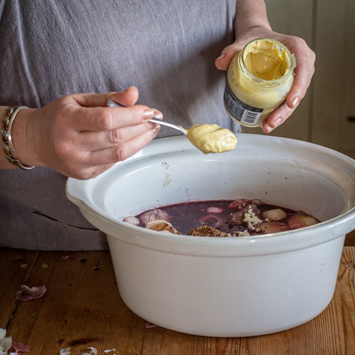 womans hands spooning dijon mustard into a white slow cooker on a wooden counter