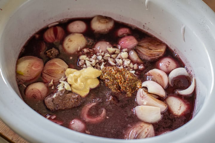 inside of a white slow cooker bowl filled with the red wine, beef chucks, shallots, mustard and garlic