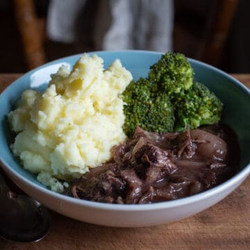 pale blue bowl on wooden kitchen surface filled with mashed potatoes, broccoli and beef in red wine stew