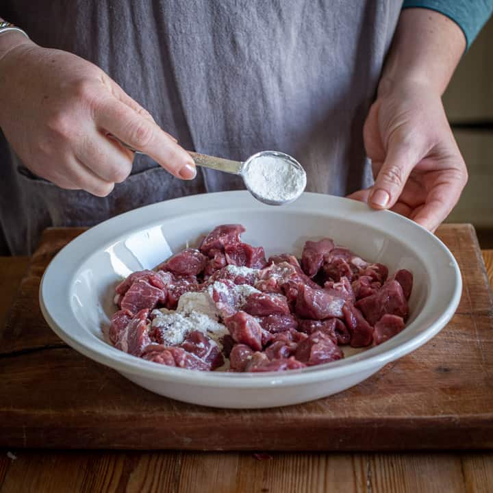 womans hands spooning flour into a white bowl of raw beef on a wooden kitchen worktop