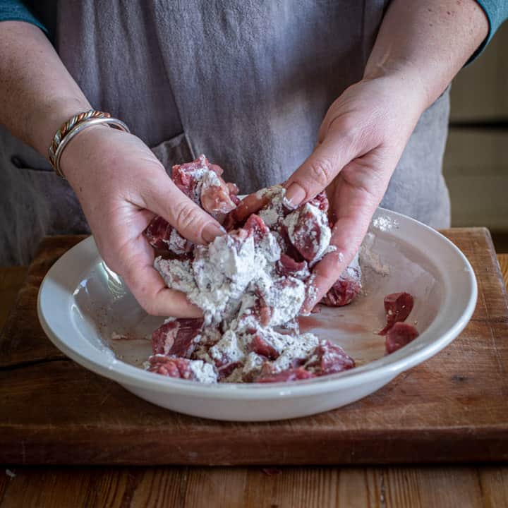 womans hands tossing raw beef chucks in flour before placing in a slow cooker