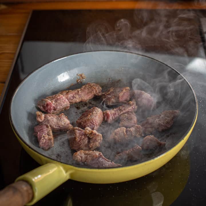 yellow enamel frying pan on a black stove top with pieces of beef searing