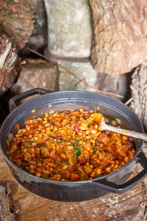 Large pan with handles sat on a wooden log containing cowboy stew recipe and a wooden spoon