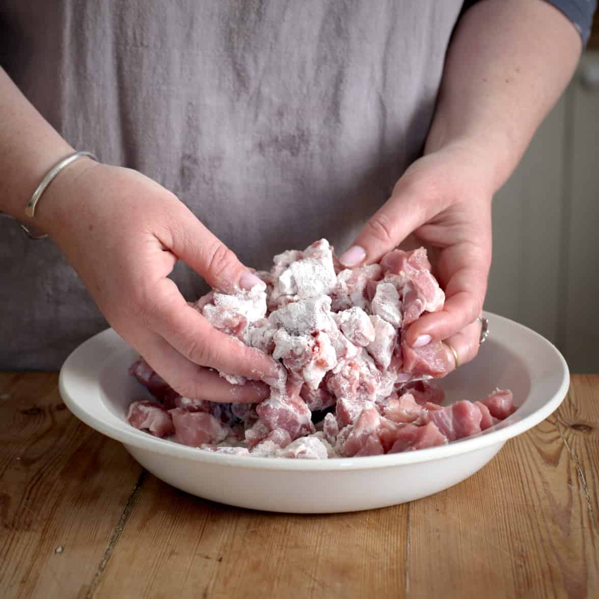 Womans hands dusting chopped pieces of pork in flour inside a flat white bowl