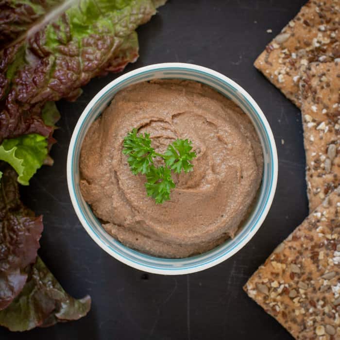 small white bowl with liver pate inside, surrounded by lettuce leaves and crackers, on a matte black surface