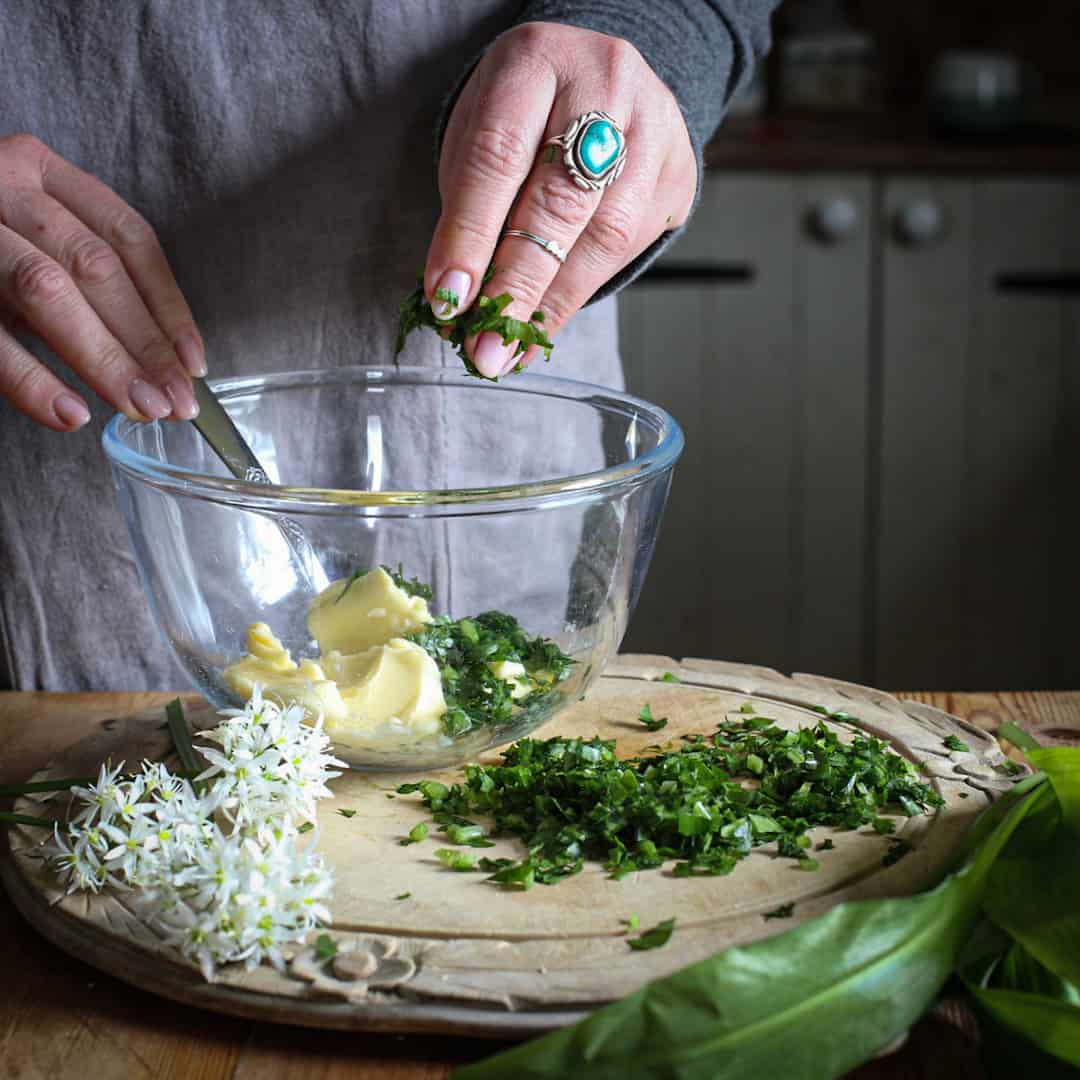 Womans hands adding chopped bright green wild garlic into a glass bowl of butter on a rustic wooden kitchen counter