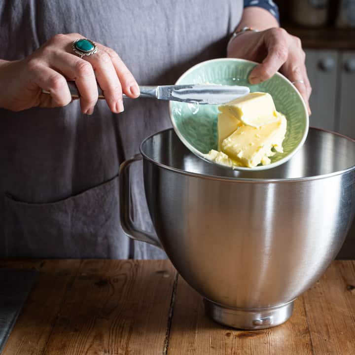 woman in grey tipping butter from a small green bowl into a silver mixing bowl