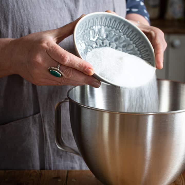 womans hands pouring white sugar from a grey bowl into a large silver mixing bowl