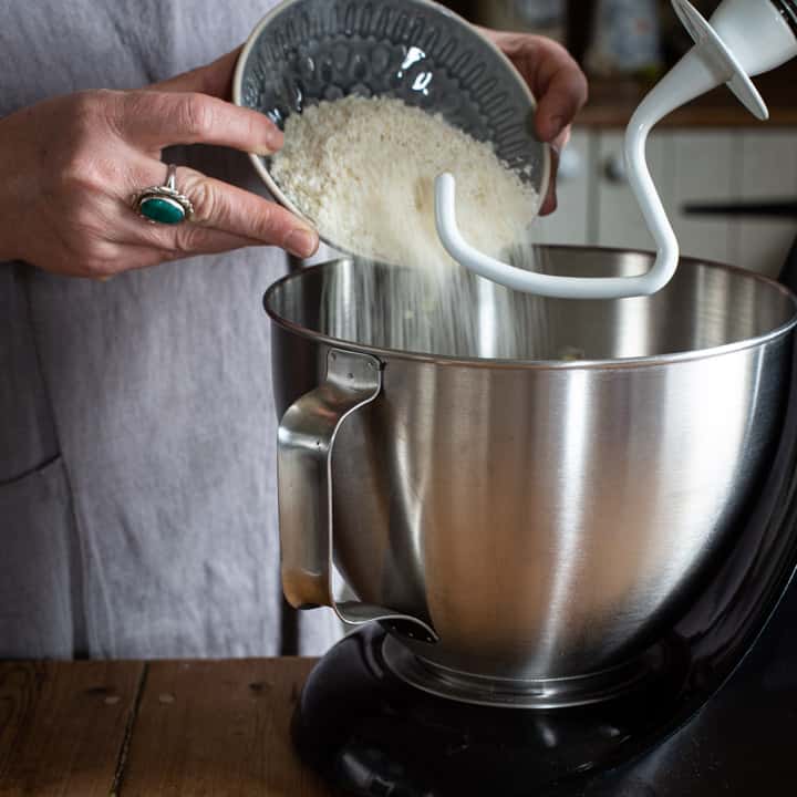 womans hands pouring desiccated coconut into a large silver mixer bowl