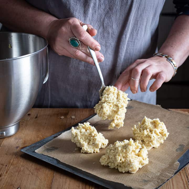 womans hands shaping coconut rock cake dough onto a silver baking sheet lined with brown greaseproof paper