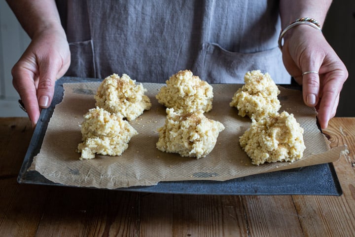 woman in grey holding a baking tray with six mounds of rock cake dough