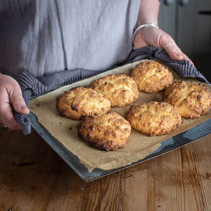 woman holding a hot silver baking tray with a grey tea towel with six golden rock cakes on