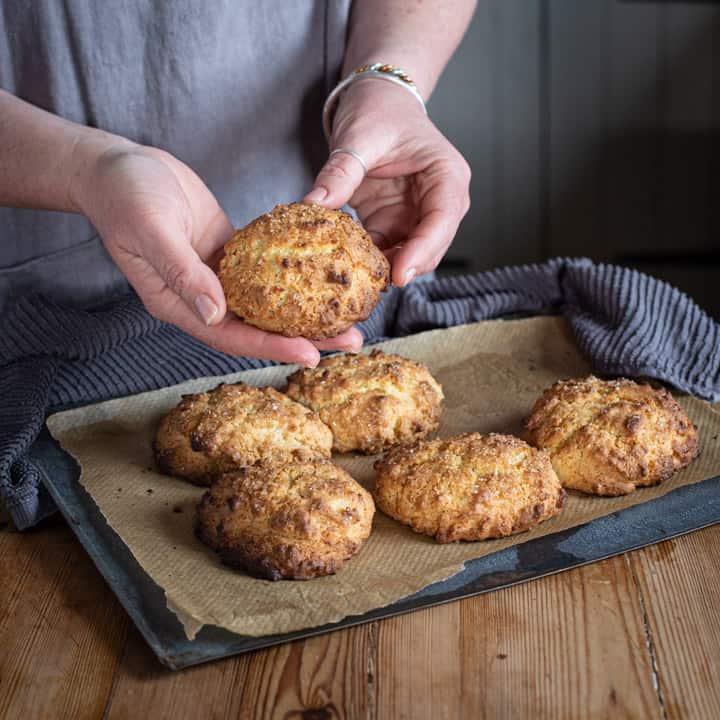 wooden kitchen counter with tray of fresh baked rock cakes and woman holding one rock cake in her hands