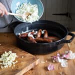 woamns hands tipping chopped onions from a blue bowl into a black cast iron pan of cooked sausages