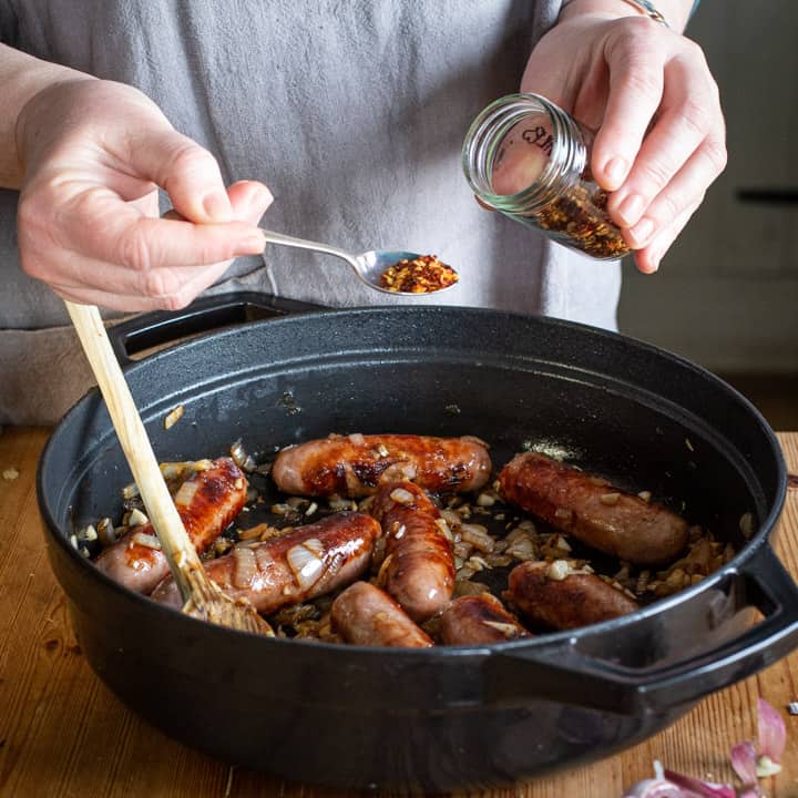 Womans hands spooning chilli flakes from a small glass spice jar into a black cast iron pan of cowboy casserole