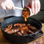 womans hands spooning baked beans from a can into a pan of sausages for a campfire stew