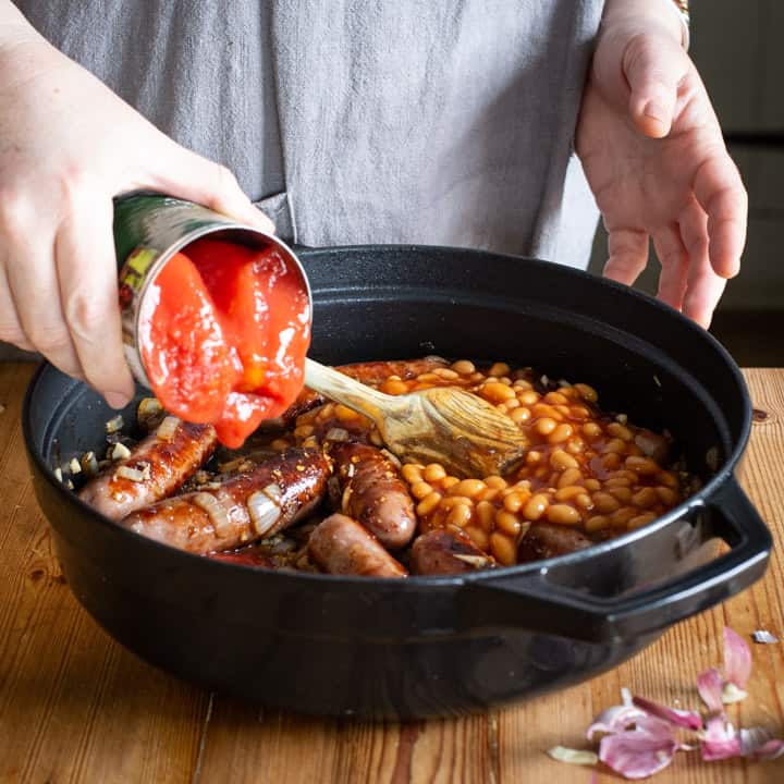 womans hands tipping a can of tomatoes into a black pan of sausage stew