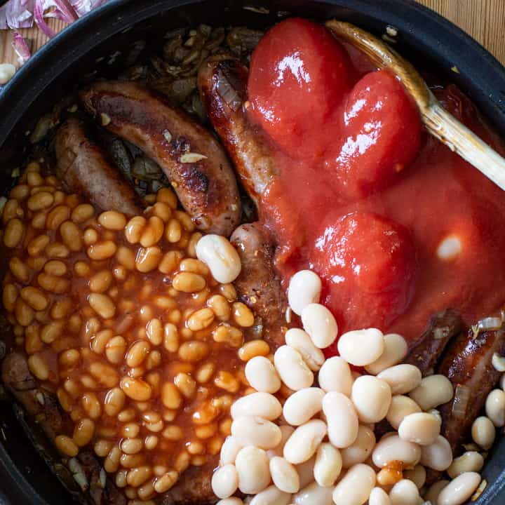 overhead shot of a black pan filled with cooked sausages, canned tomatoes, white beans and baked beans