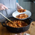 womans hands dishing up cowboy sausage casserole from a black cast iron pan into a white enamel camping plate