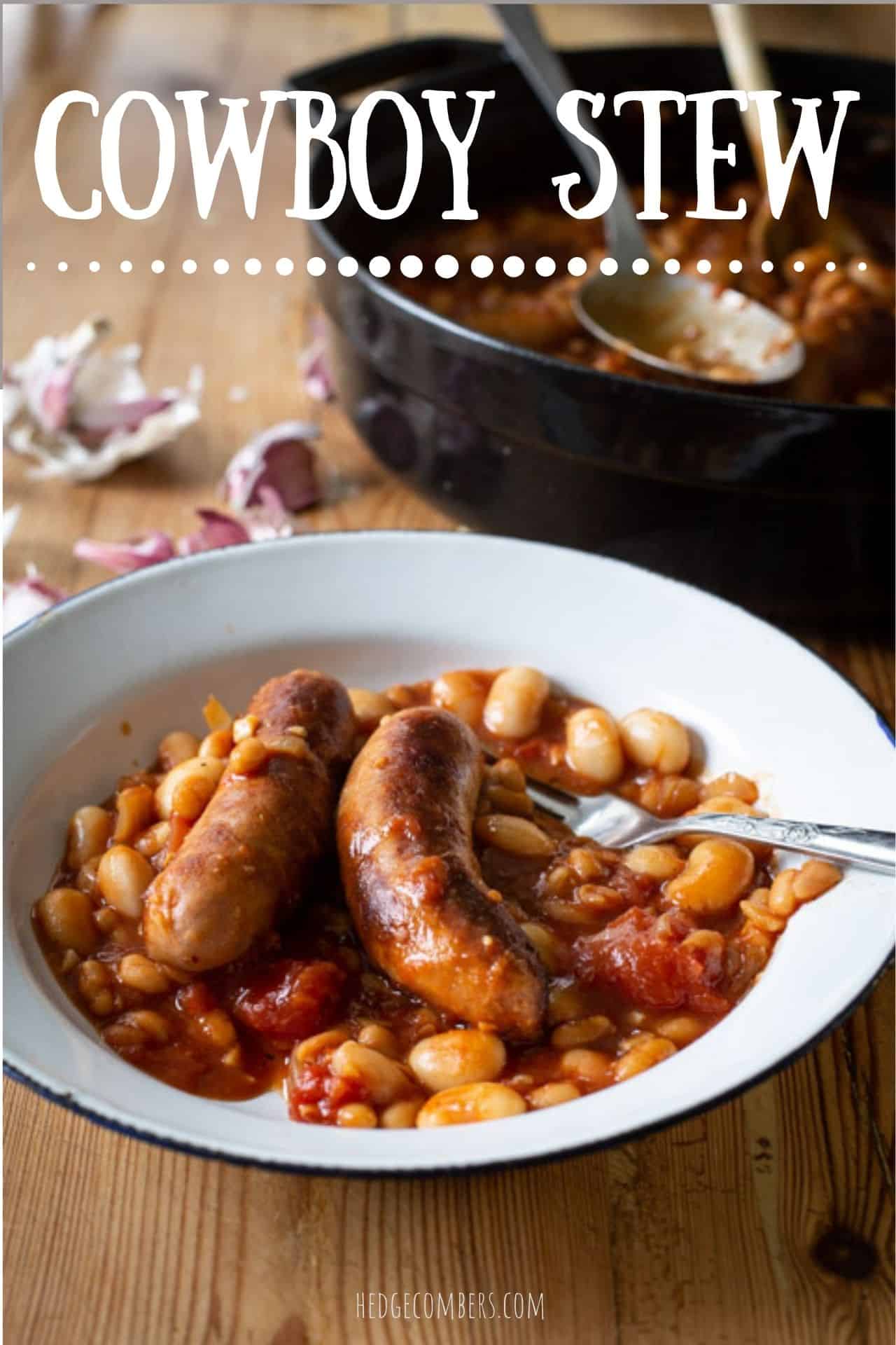 white enamel bowl of sausage and bean cowboy stew on a wooden counter with a black cooking pan and garlic cloves in the background