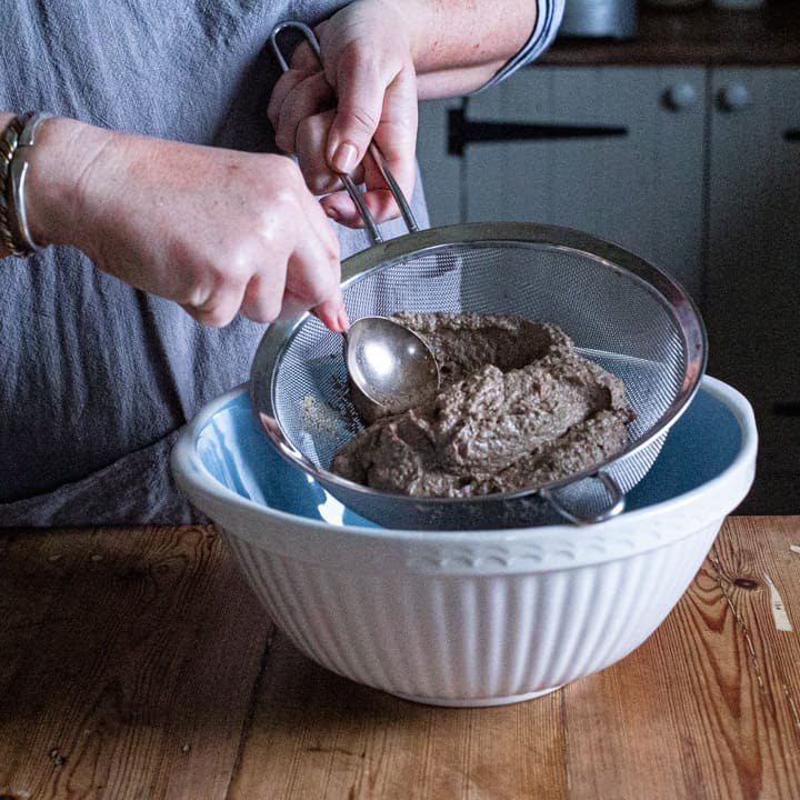 wooden kitchen counter with large blue and white bowl and womans hands pushing fresh homemade pate through a metal sieve with a soup spoon 