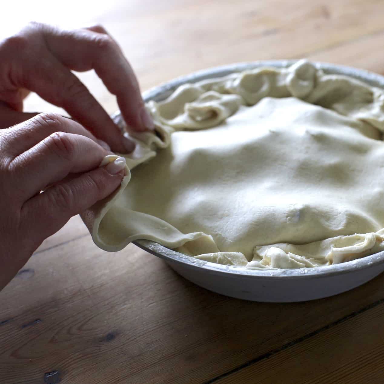 Womans hands tucking in a sheet of puff pastry to a silver tarte tatin pie dish filled with rhubarb pieces