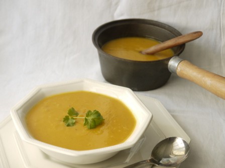 Sweet Potato and Garlic Soup in a white bowl on a white plate with a soup spoon beside a pan of soup with a wooden spoon all on a white cloth