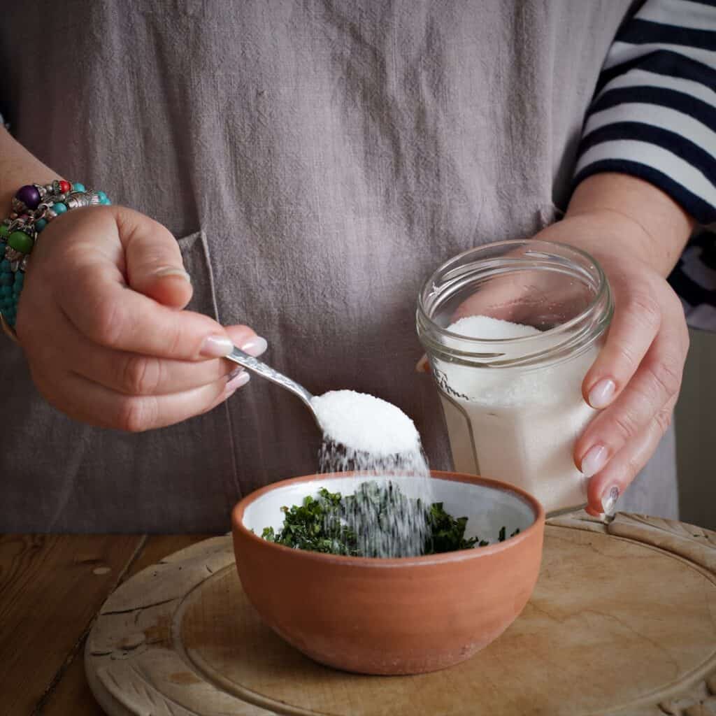 Woman in grey scattering white sugar from a silver teaspoon over a small brown bowl of fresh mint sauce ingredients