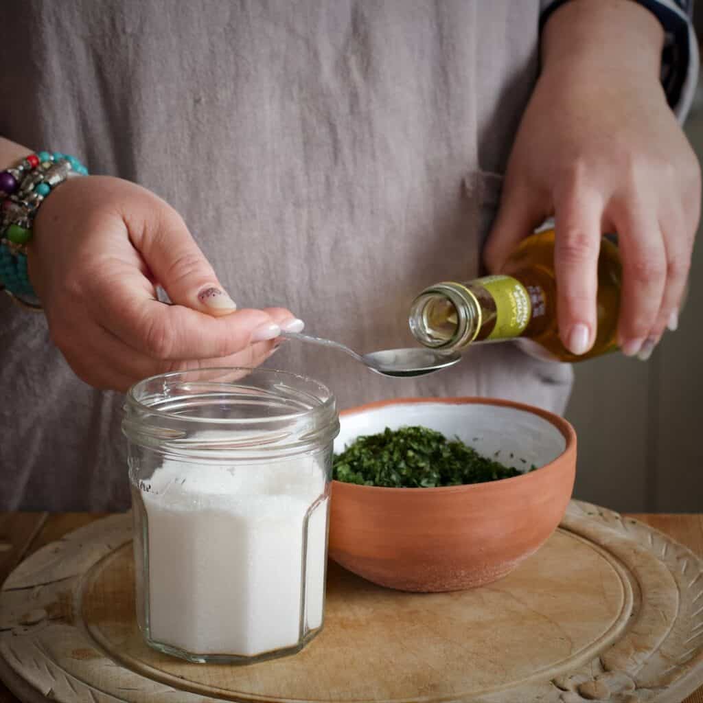 Womans hands measuring out apple cider vinegar from a glass bottle into a teaspoon over a terracotta bowl of mint leaves