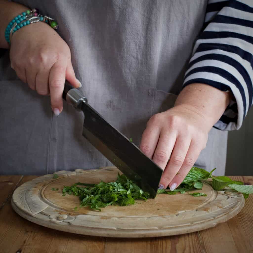 Woman in grey finely chopping mint with a large Japanese knife on a rustic wooden chopping board