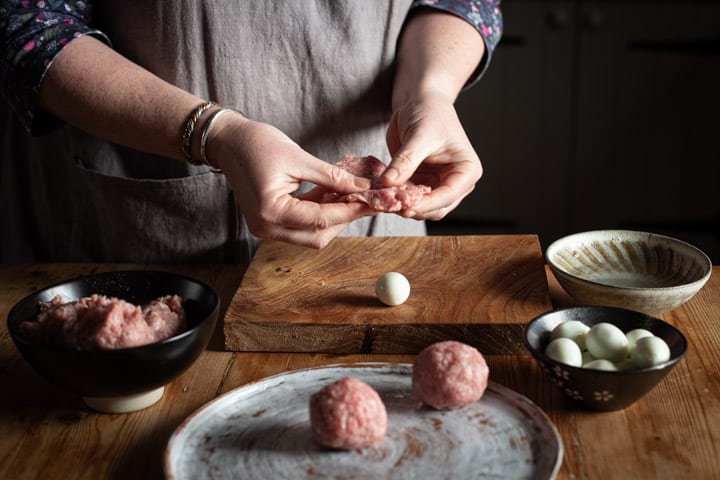 womans hands covering qualis eggs with sausagemeat to make mini scotch eggs in a rustic wooden kitchen setting