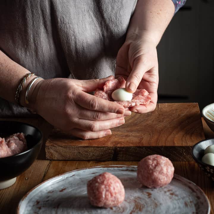 womans hands shaping sausagemeat around a hard boiled quails egg to make mini scotch eggs over a wooden kitchen counter