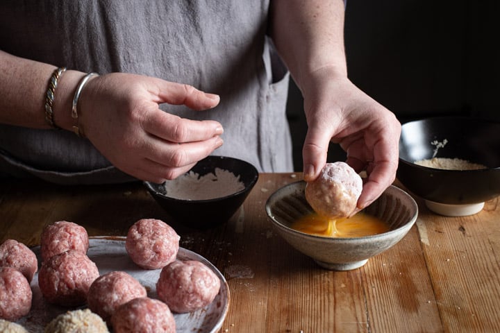 womans hands dipping a scotch egg into beaten egg before rolling in breadcrumbs