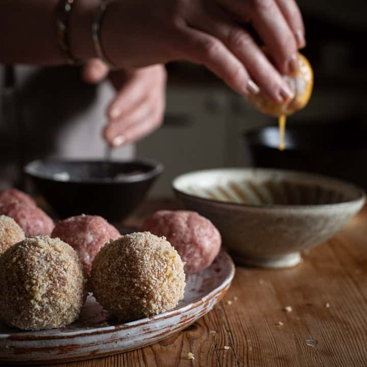 wooden kitchen counter and tray of scotch eggs being made with hands in background lifting a scotch egg out of eggwash