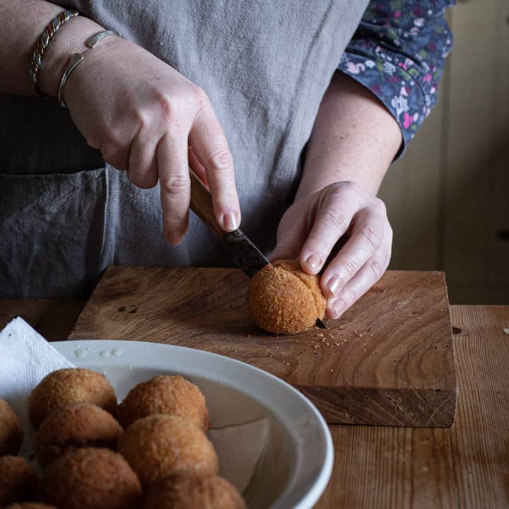 womans hands cutting a scotch egg in half on a wooden chopping board with white bowl of more eggs close by