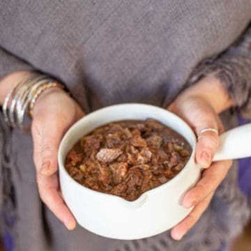 womans hands holding white pot of beef and ale stew against a grey jumper