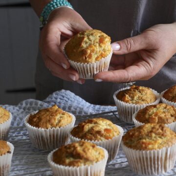 Womans hands holding a warm apricot muffin over a cooling rack of serveral more
