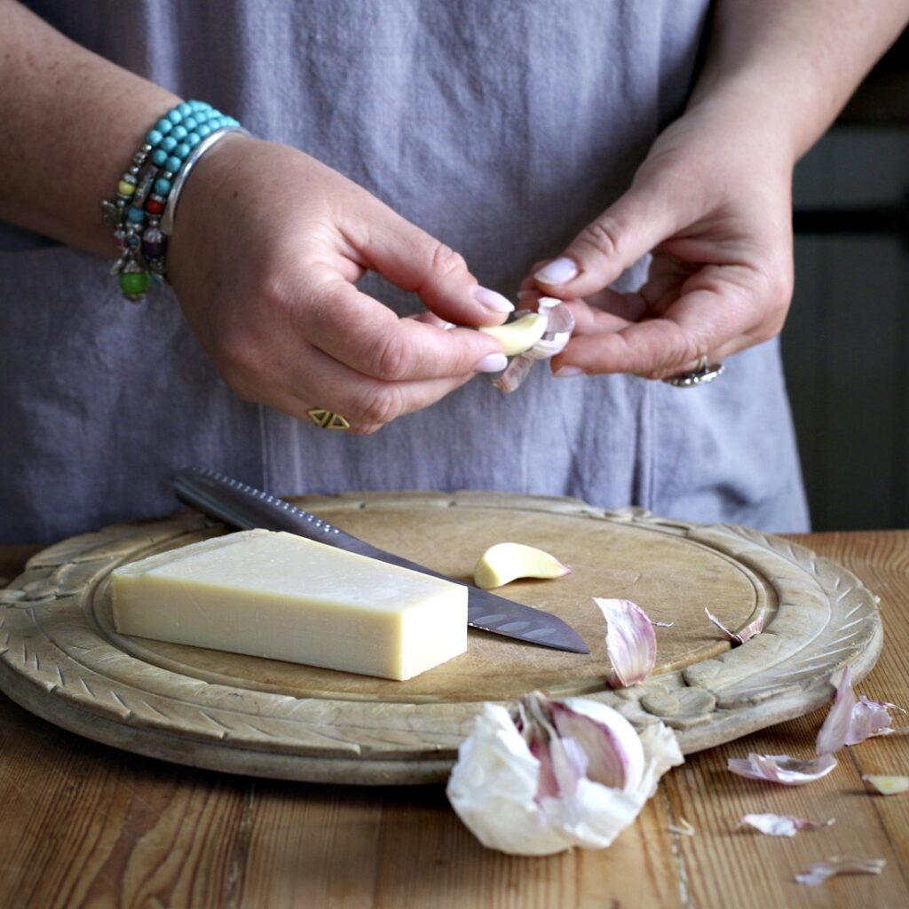 Womans hands holding a garlic clove over an old fashioned wooden chopping board holding more garlic and a block of Parmesan cheese